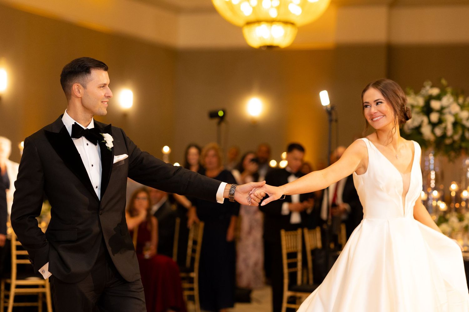 A couple performs their first dance surrounded by guests in an elegant ballroom with floral decorations.