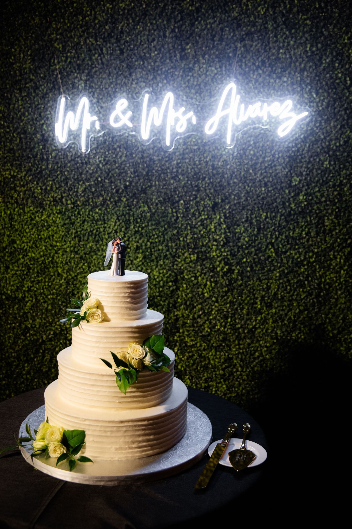 White tiered wedding cake with neon sign reading 'Mr & Mrs' against green hedge backdrop.