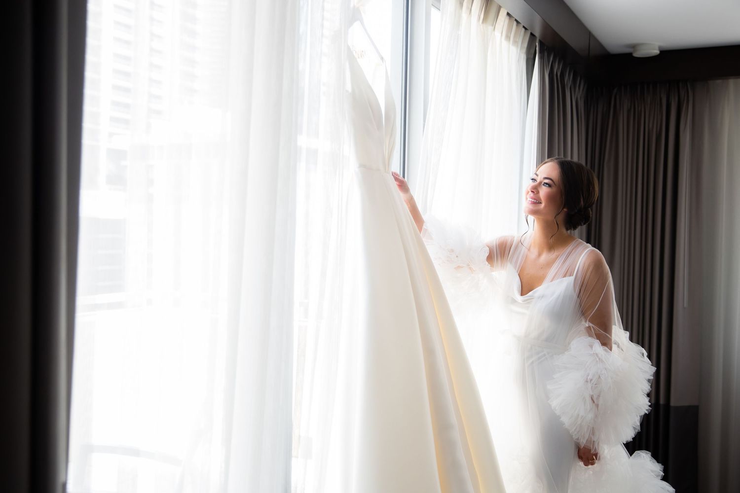 Wedding dress silhouette captured against bright window with flowing curtains.