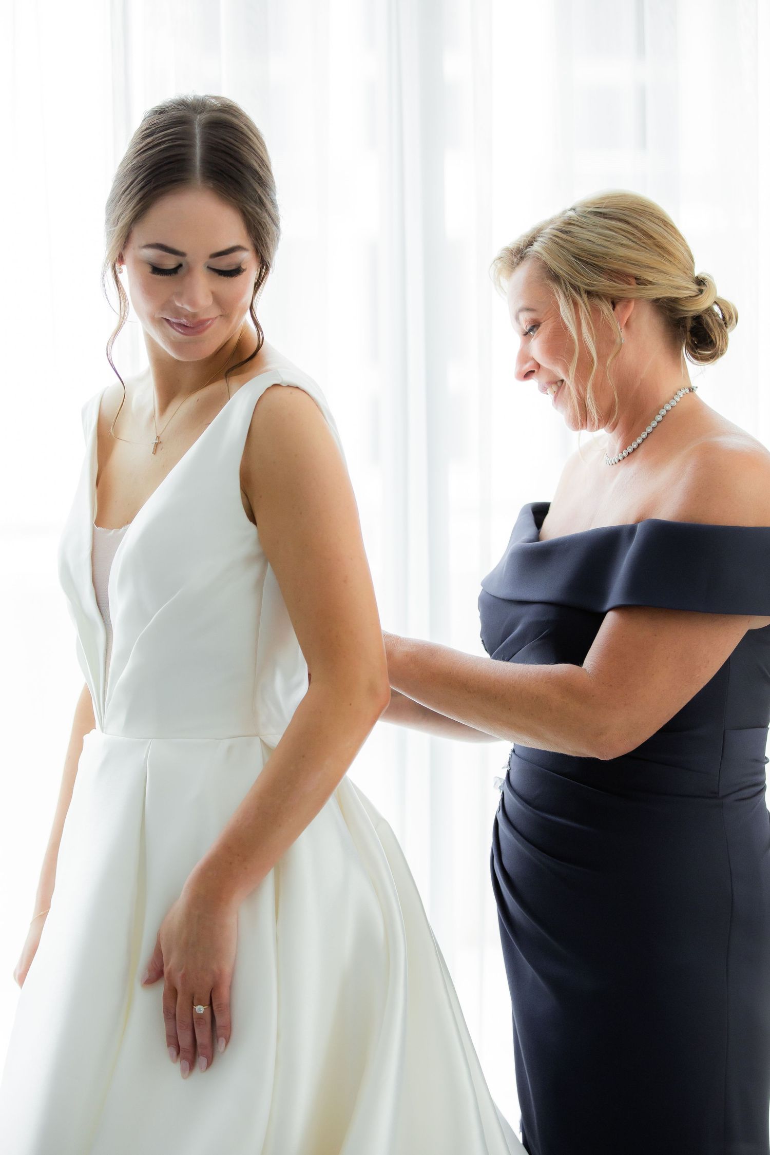 Bride and bridesmaid in navy dress share intimate moment during wedding preparations.