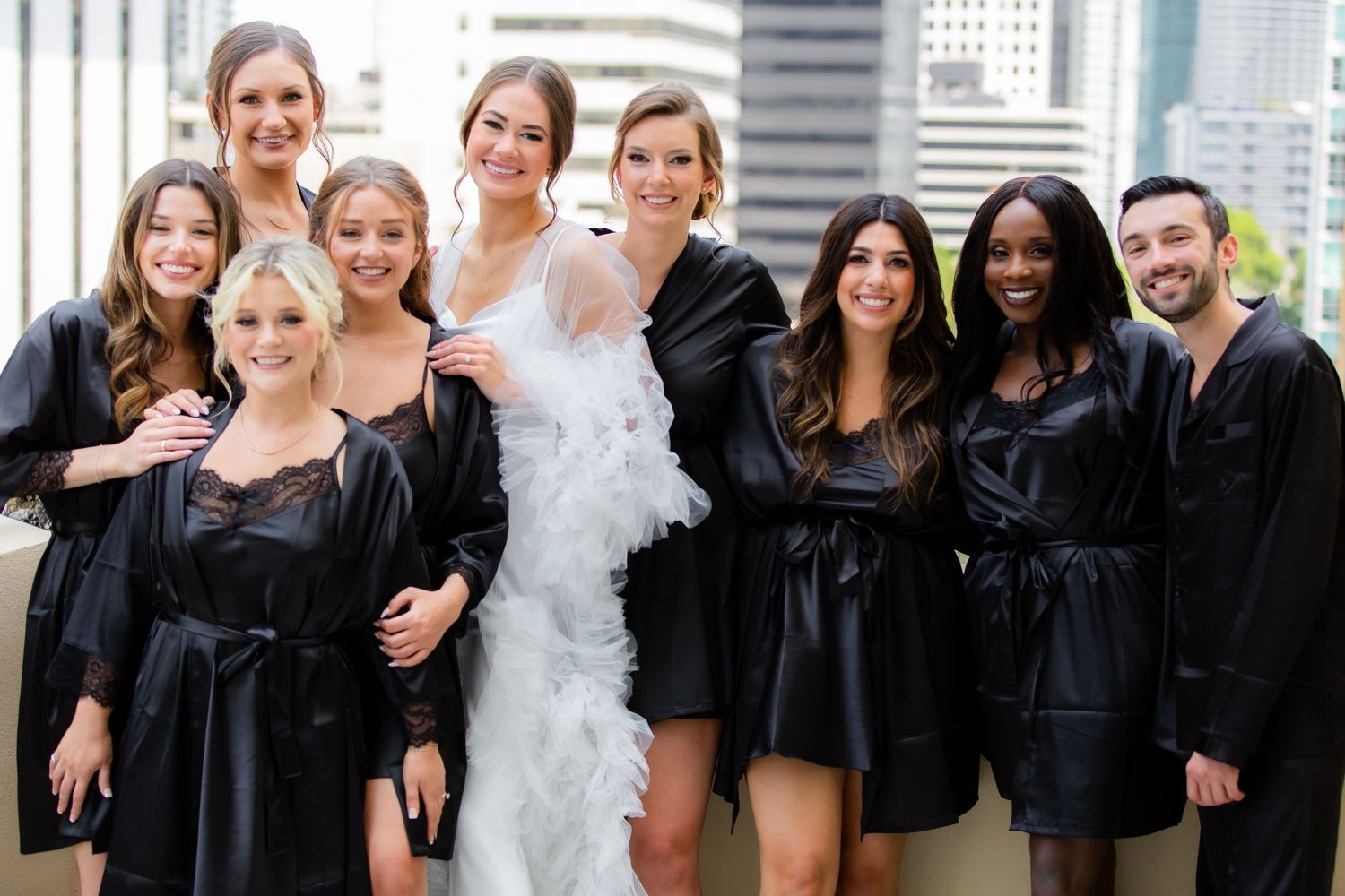 Wedding party in black attire poses together on a modern urban rooftop with city views.