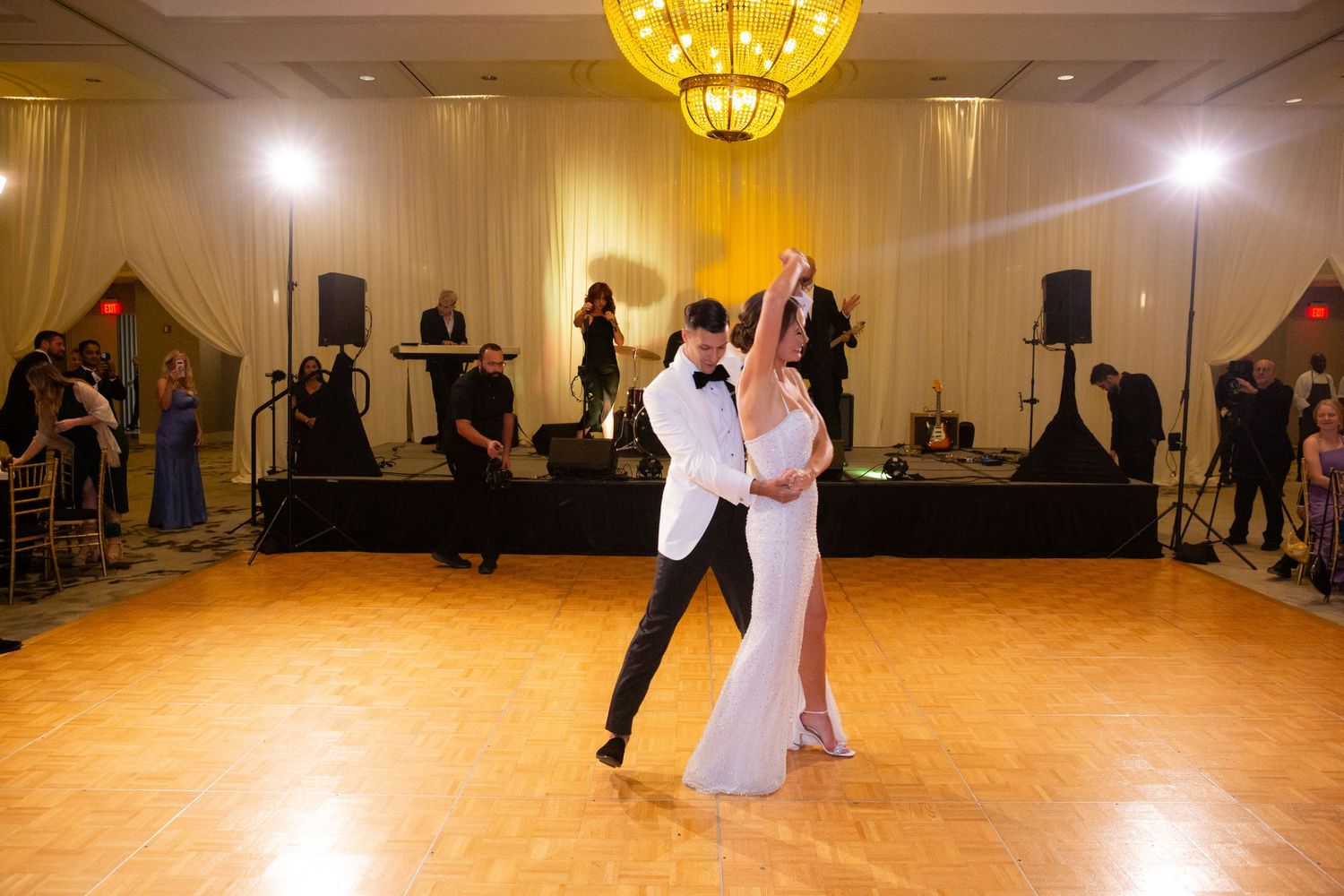 Wedding reception dance floor scene with guests dancing under golden chandeliers.