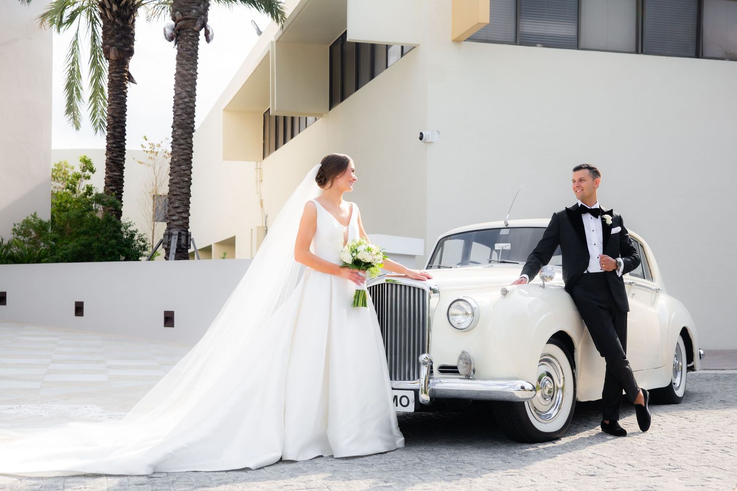 Elegant wedding couple poses next to vintage white car outside a building with palm trees.