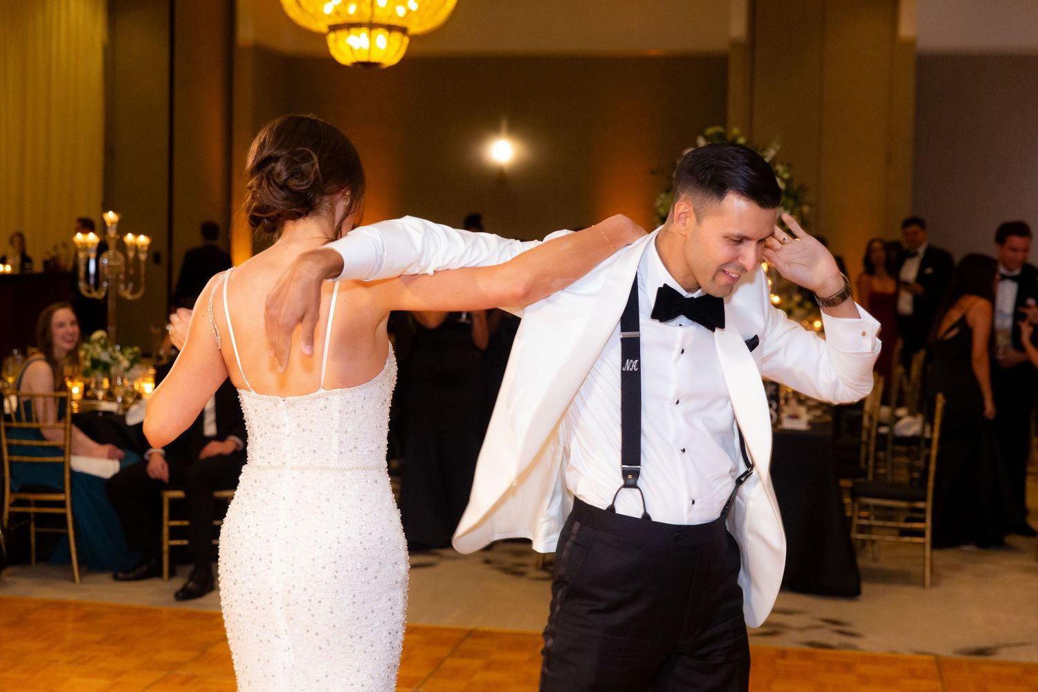 Wedding reception guests dancing and celebrating on a wooden dance floor under warm chandelier lighting.