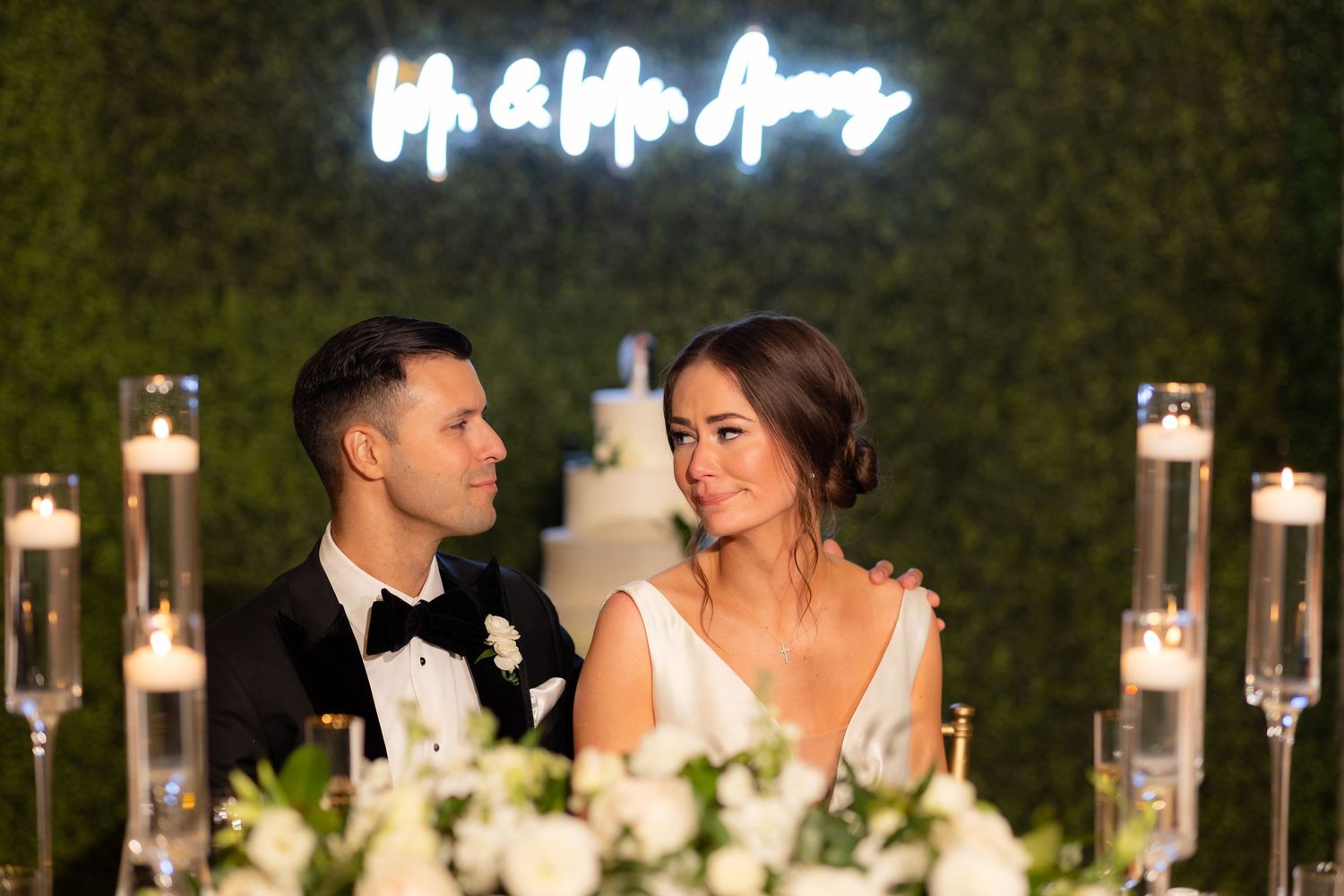 Wedding party seated at decorated head table with illuminated backdrop and candles.