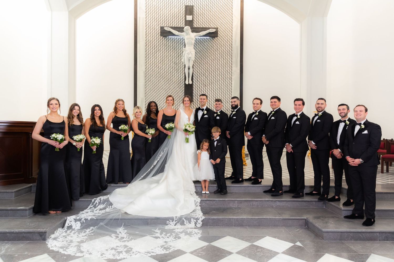 Large wedding party posed formally alongside white wall in classic black attire.