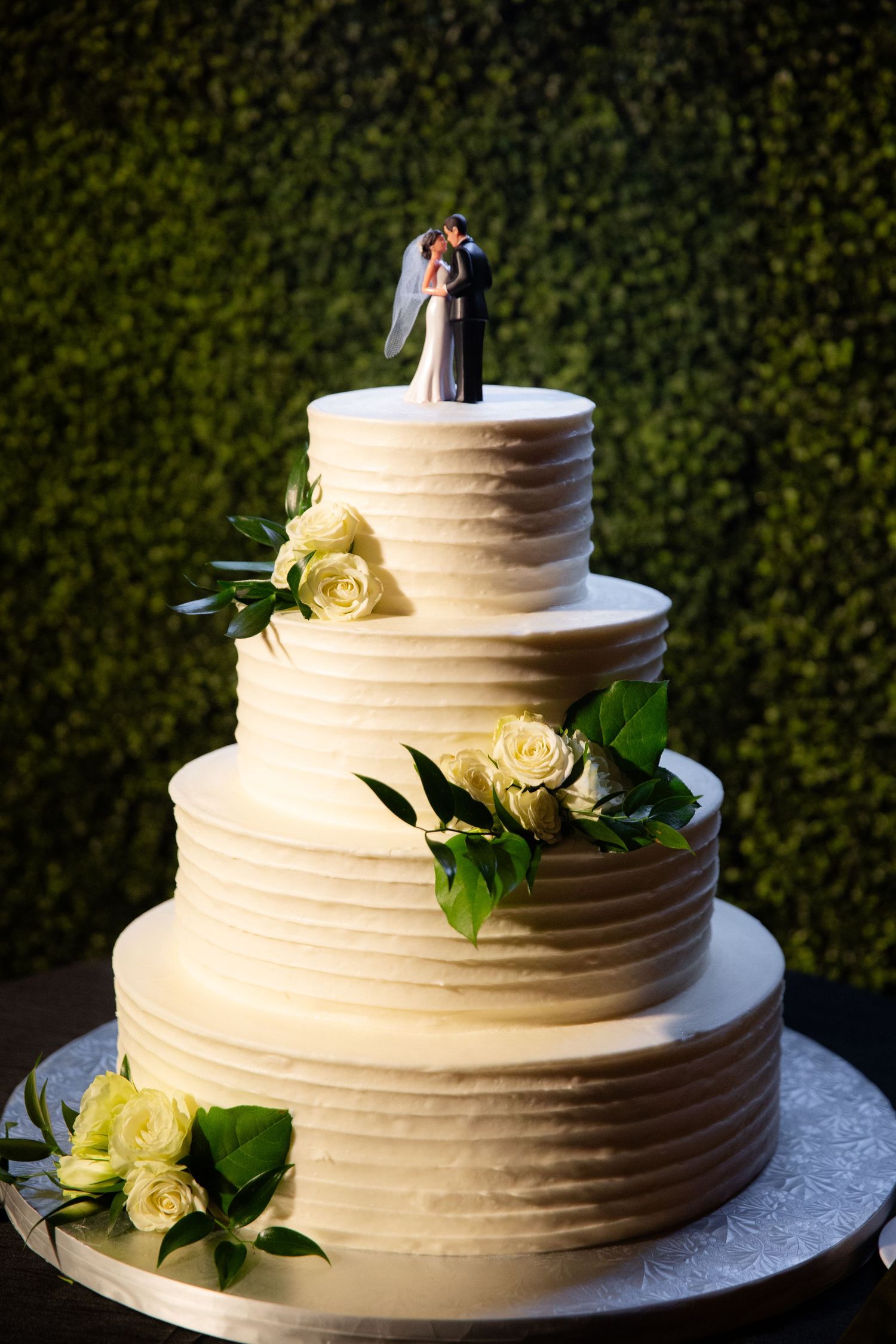 Elegant four-tier white wedding cake decorated with yellow roses and greenery against a hedge wall.