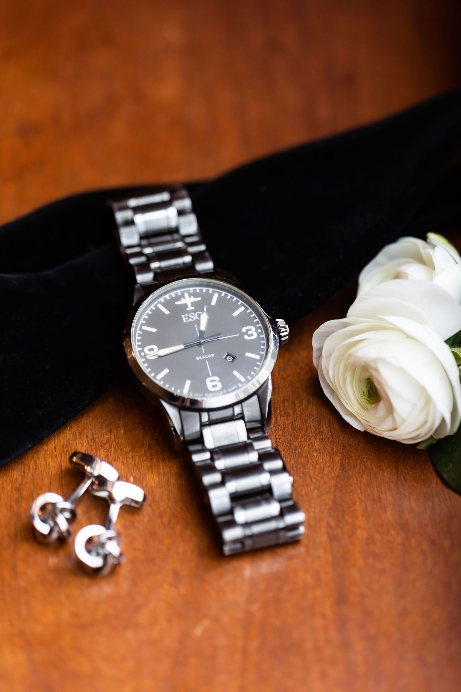 Wedding accessories including silver watch, cufflinks, and white rose on wooden surface.