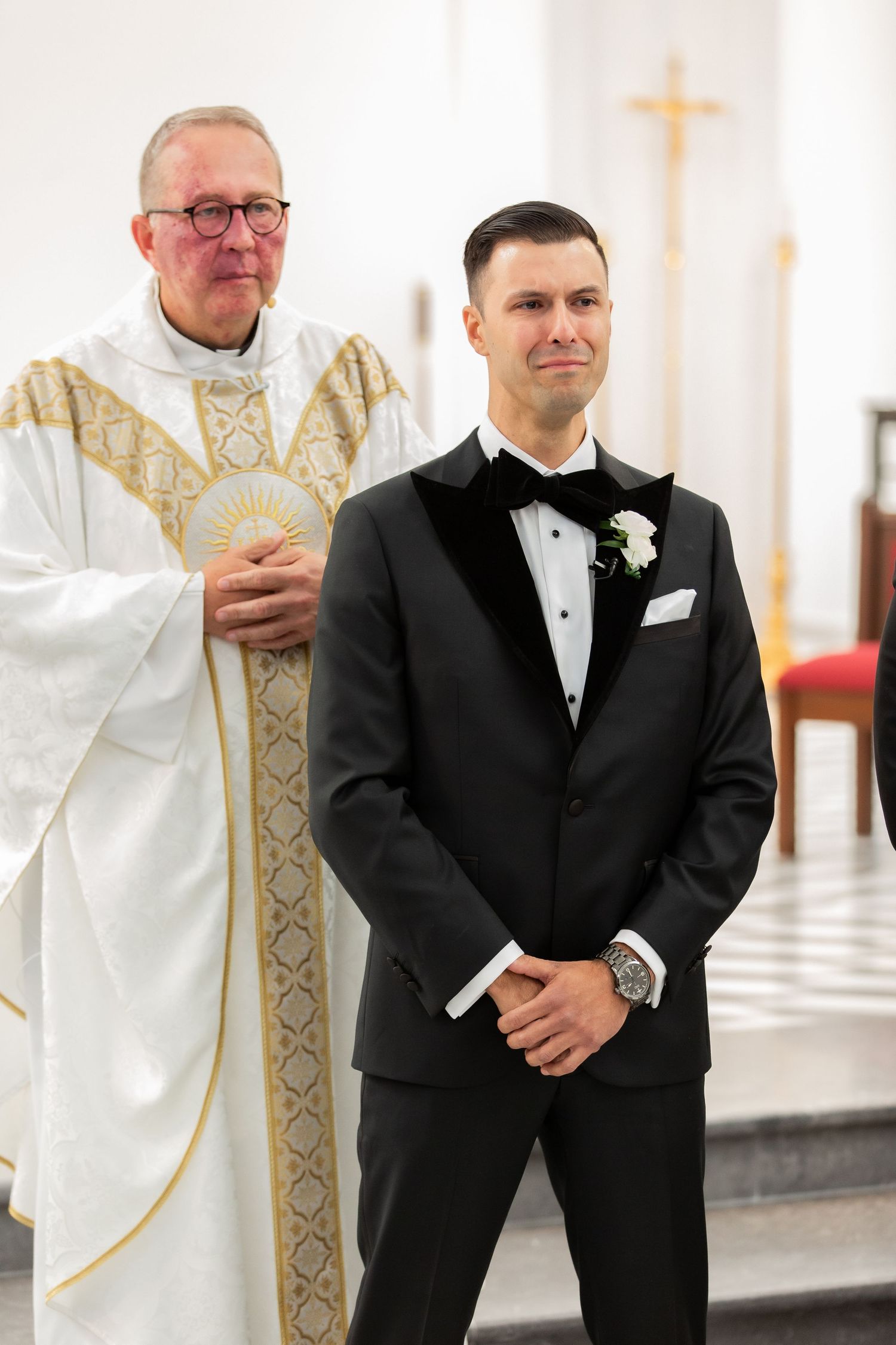 Two people stand together at a church altar, one wearing traditional white religious vestments.