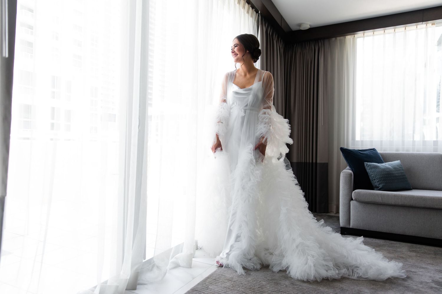 A bride in a luxurious white wedding gown poses gracefully in a modern hotel room with floor-to-ceiling windows.