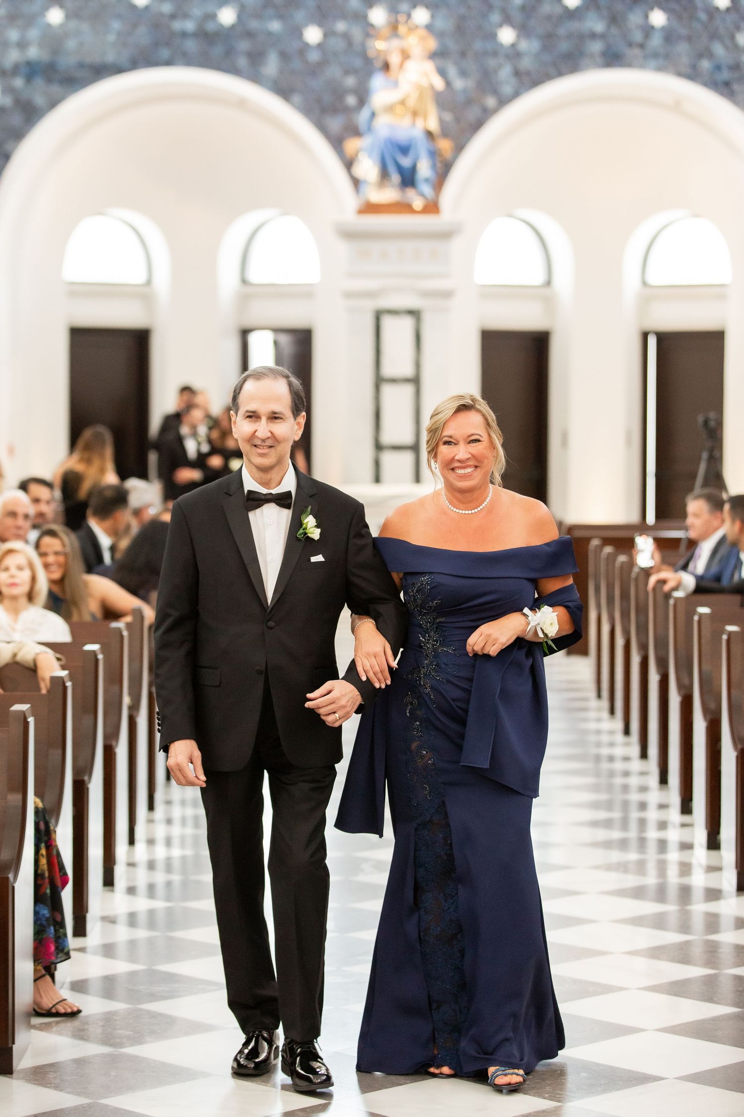 Wedding attendants in elegant black attire walking arm-in-arm down a grand church aisle.