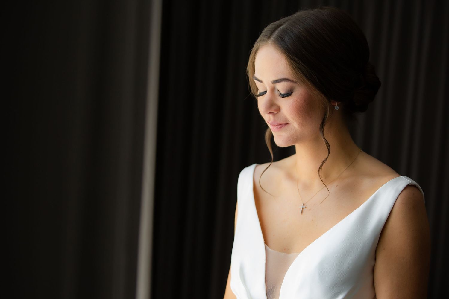 Bride in sleeveless white gown illuminated by soft window light during preparation.