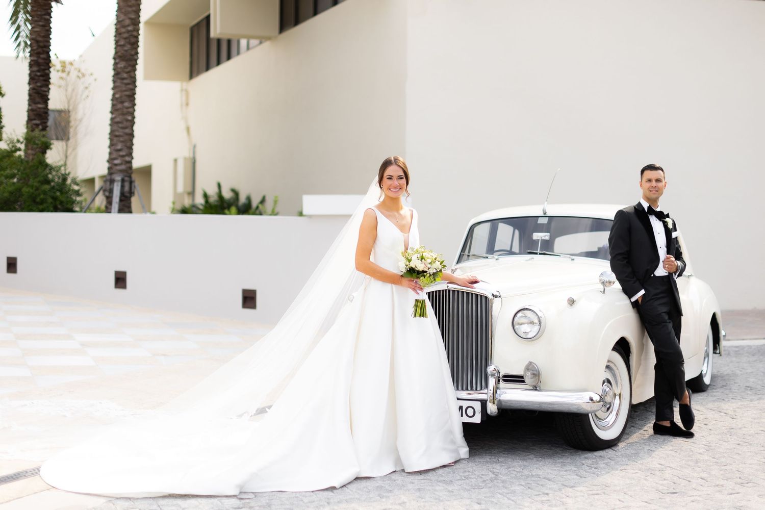 Wedding couple poses with classic white Rolls Royce at venue entrance.