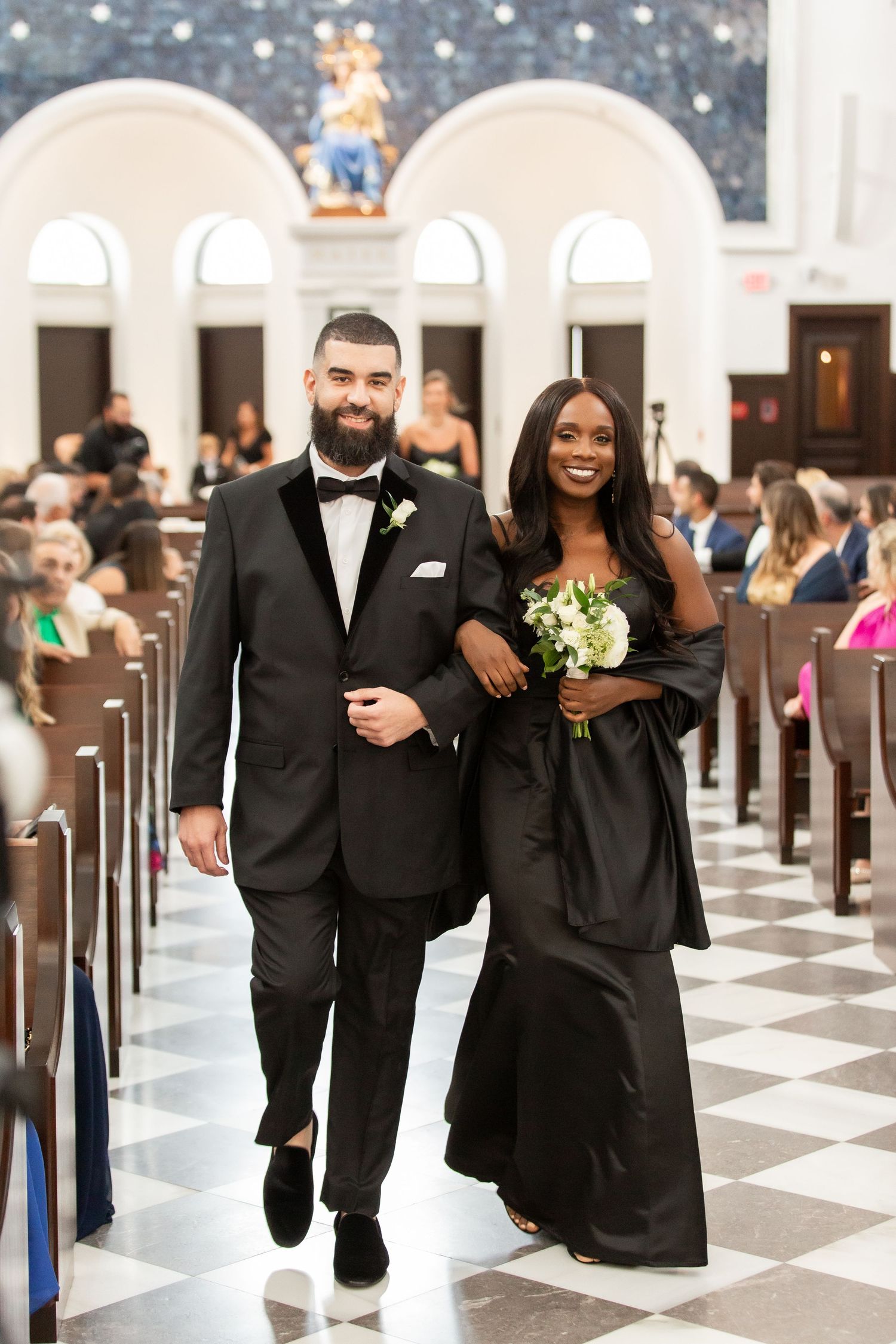 Wedding party members in elegant black attire processing down a church aisle with checkered flooring.