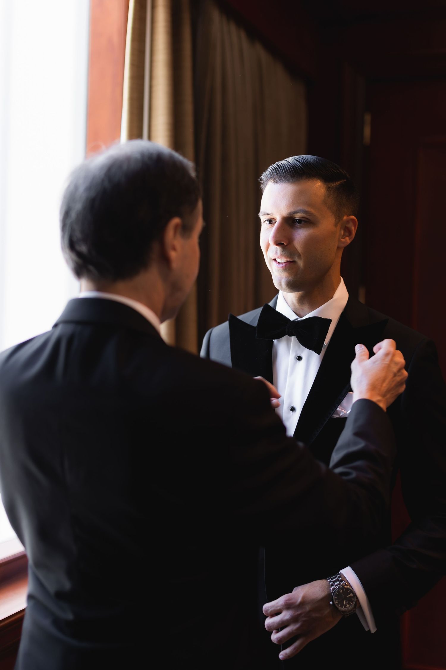 Men in formal black tuxedos share an emotional moment in a dimly lit room.