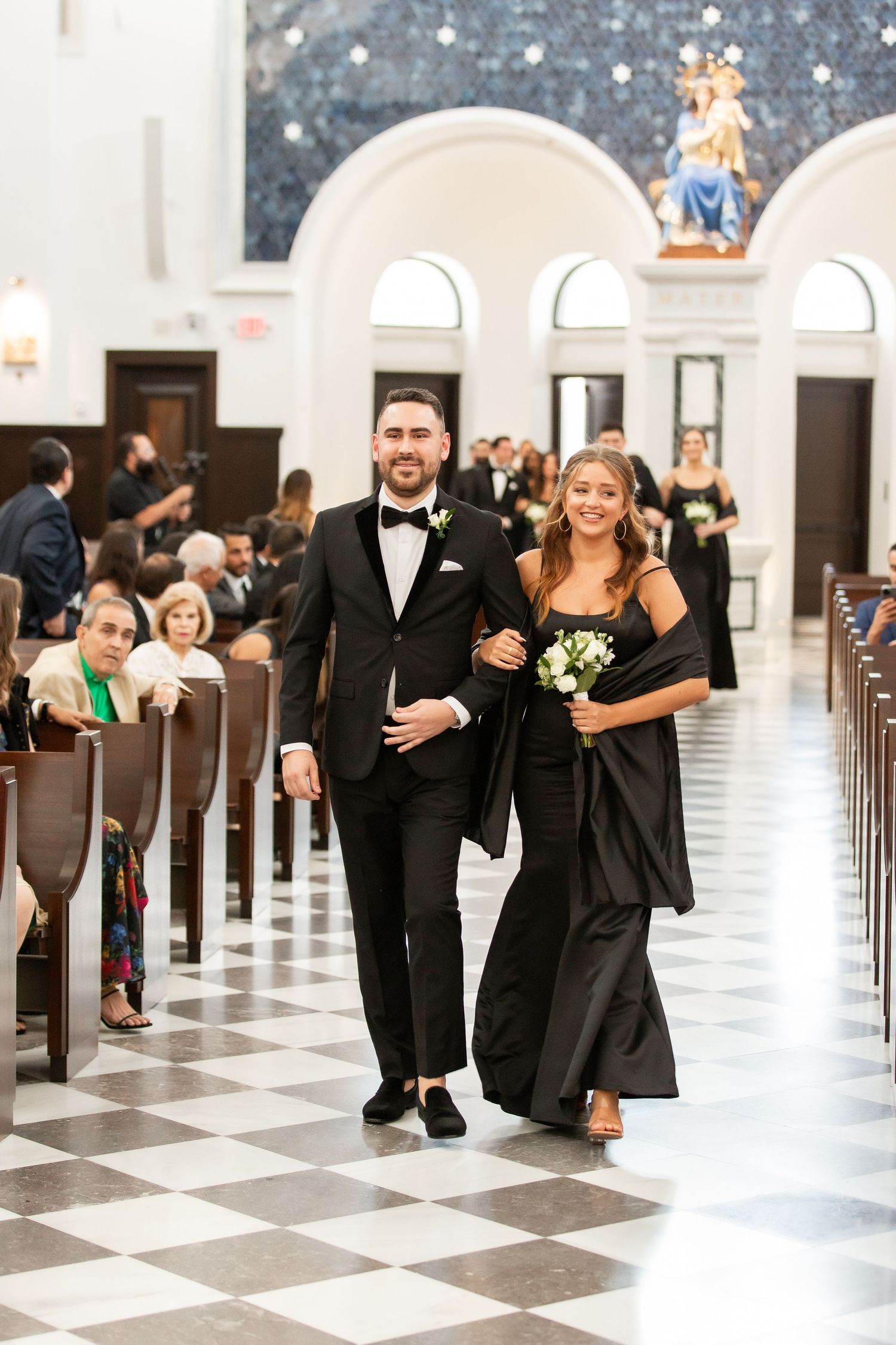 Wedding party processional with pairs walking down a checkered floor aisle in a church with arched windows.