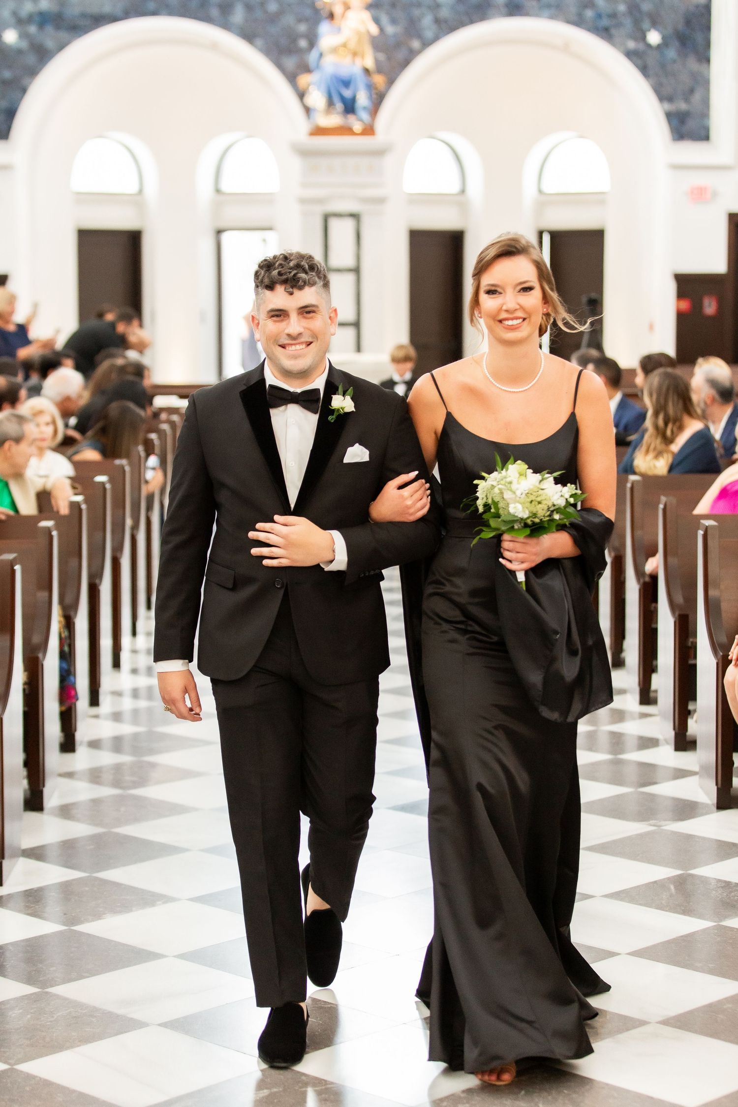 Wedding attendants in formal black attire walking arm-in-arm down a church aisle.