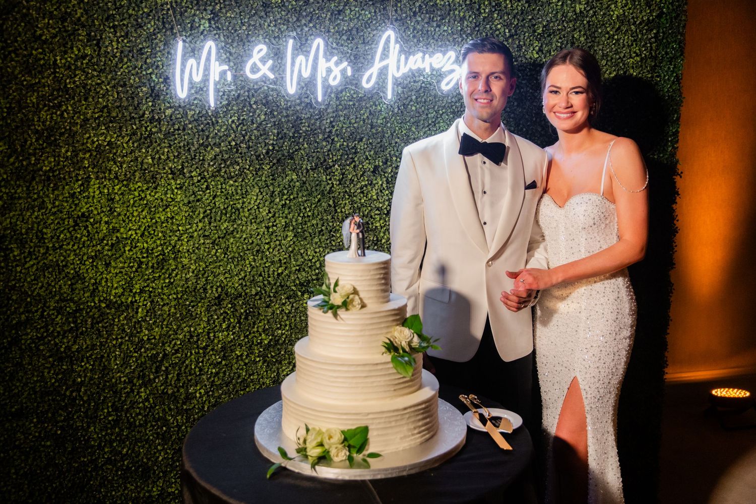 Newlyweds posing with their wedding cake under a neon sign against a green hedge wall.
