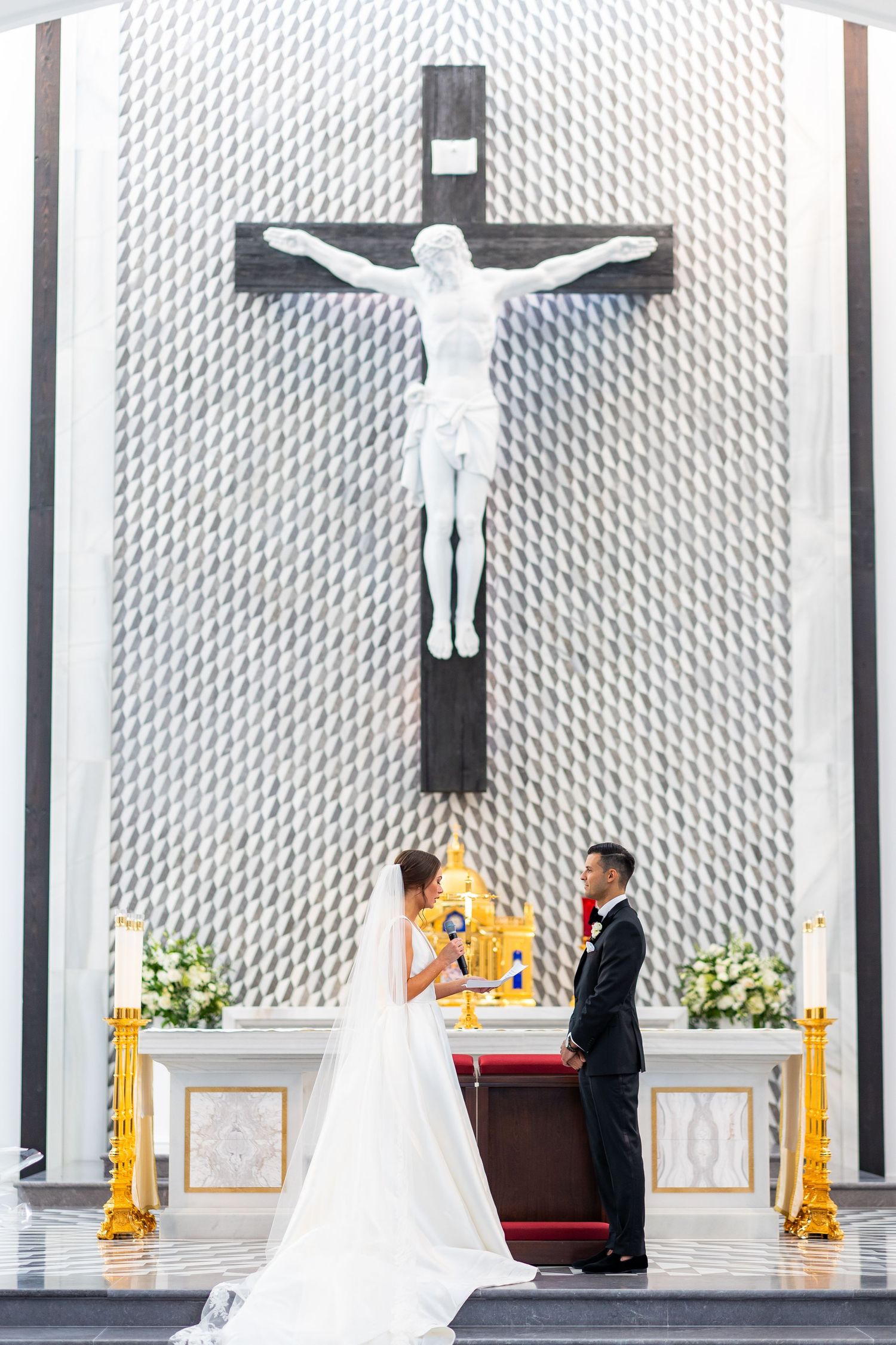 A wedding ceremony takes place in a contemporary church with a distinctive white textured wall and large crucifix.