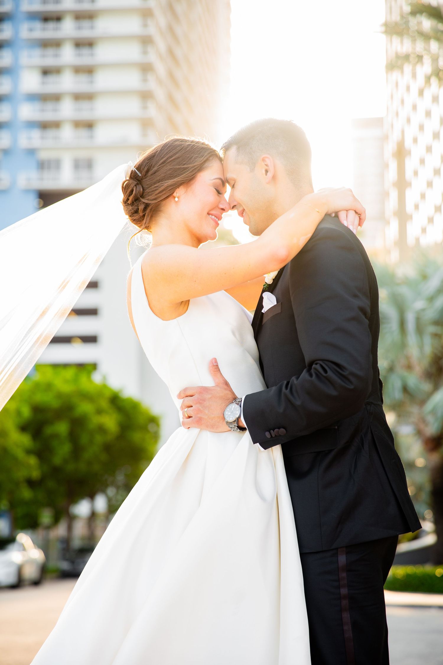 Elegant couple shares intimate moment during wedding photoshoot at sunset near luxury buildings.