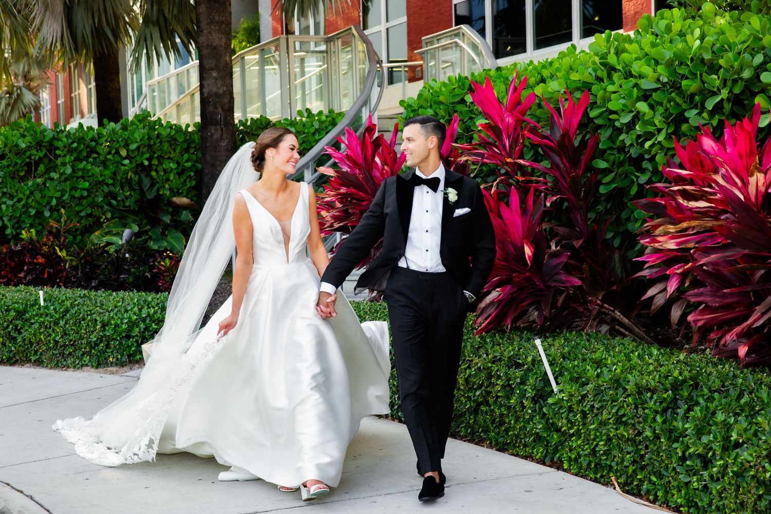 Romantic wedding couple poses in a lush tropical garden setting with vibrant red flowers and greenery.
