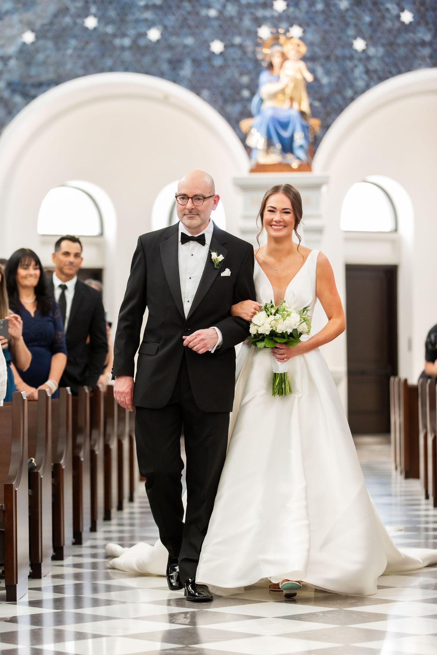 Wedding procession walking down church aisle with checkered floor and arched architecture.