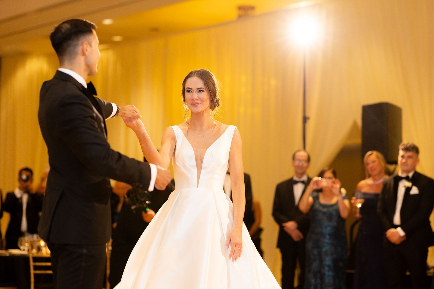 A couple shares their first dance in a beautifully lit ballroom with watching guests.
