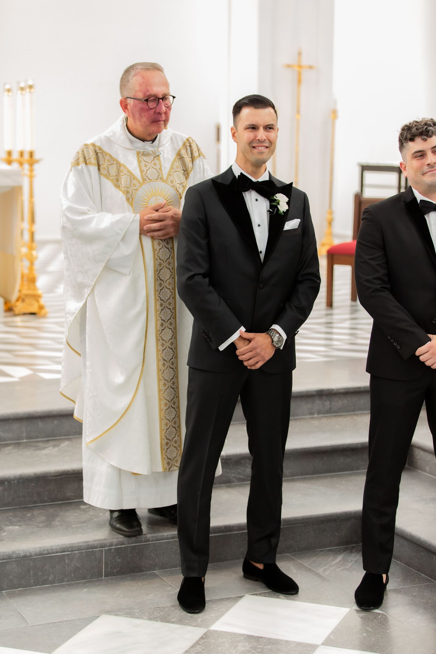 Clergy member in white robes with gold embroidery stands next to a person in formal black attire in a church setting.
