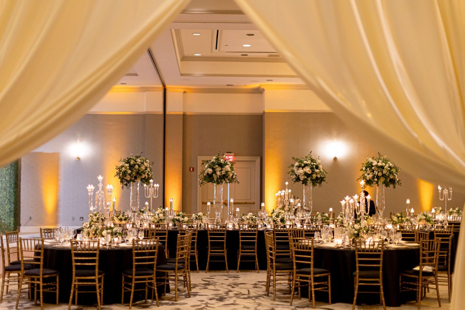 Elegant ballroom wedding reception setup with white draping, chandeliers and round tables.