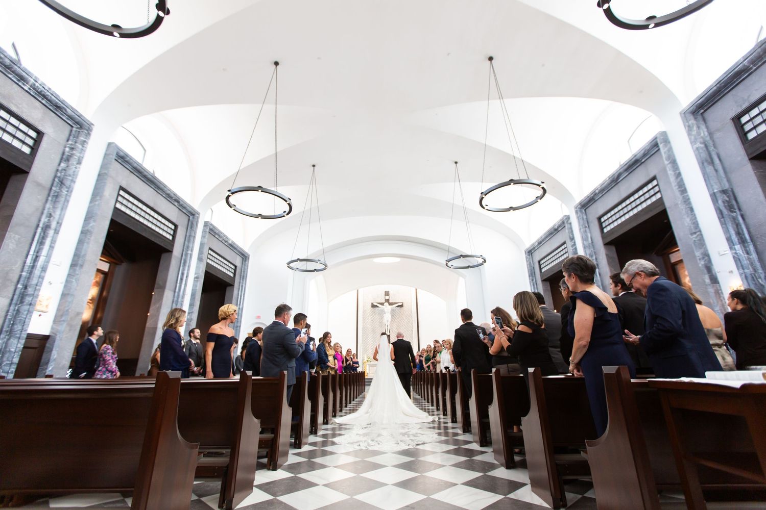 Church interior with rows of wooden pews leading to a bright white altar under vaulted ceilings.