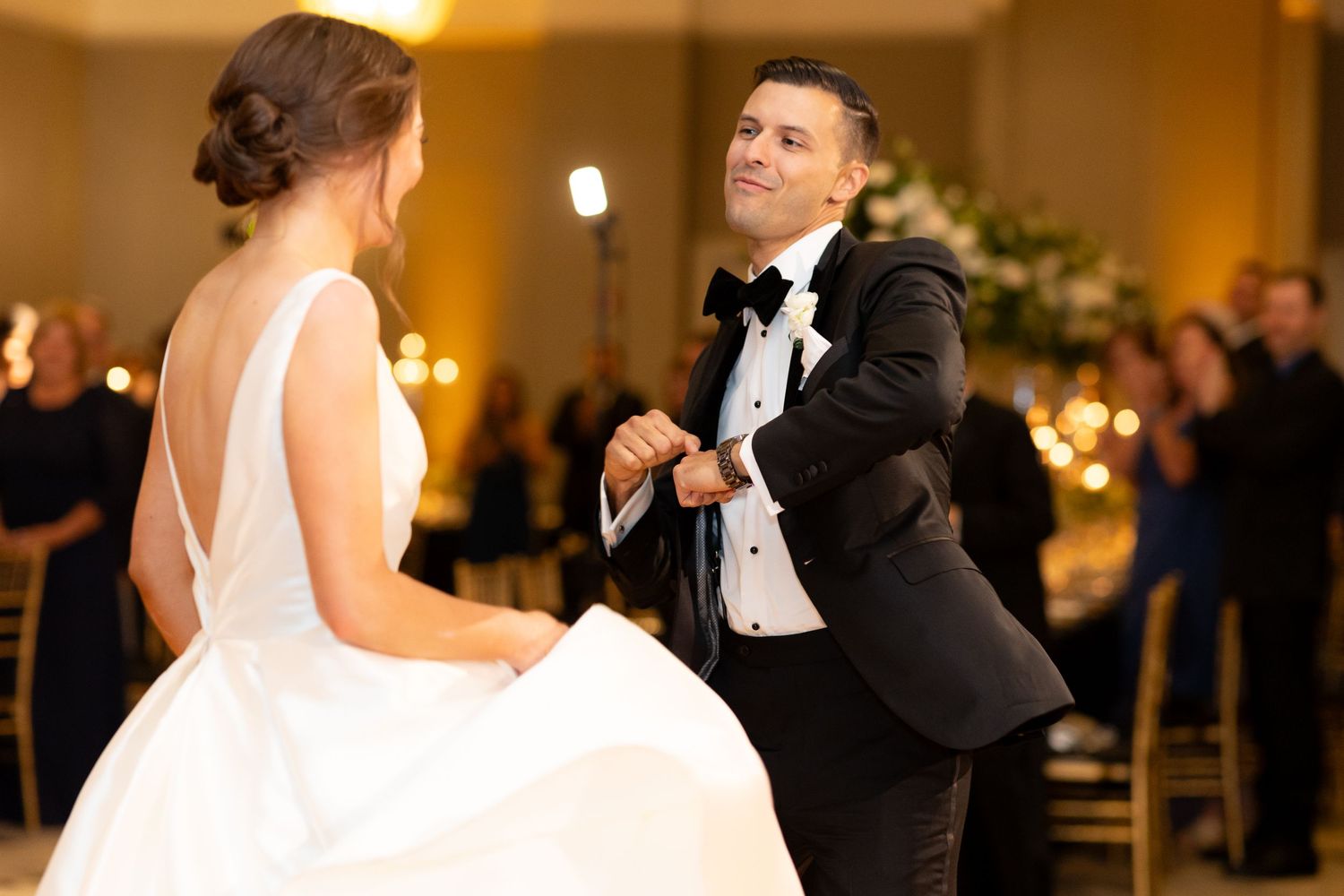 A couple shares an energetic first dance at their wedding reception in an elegantly lit ballroom.