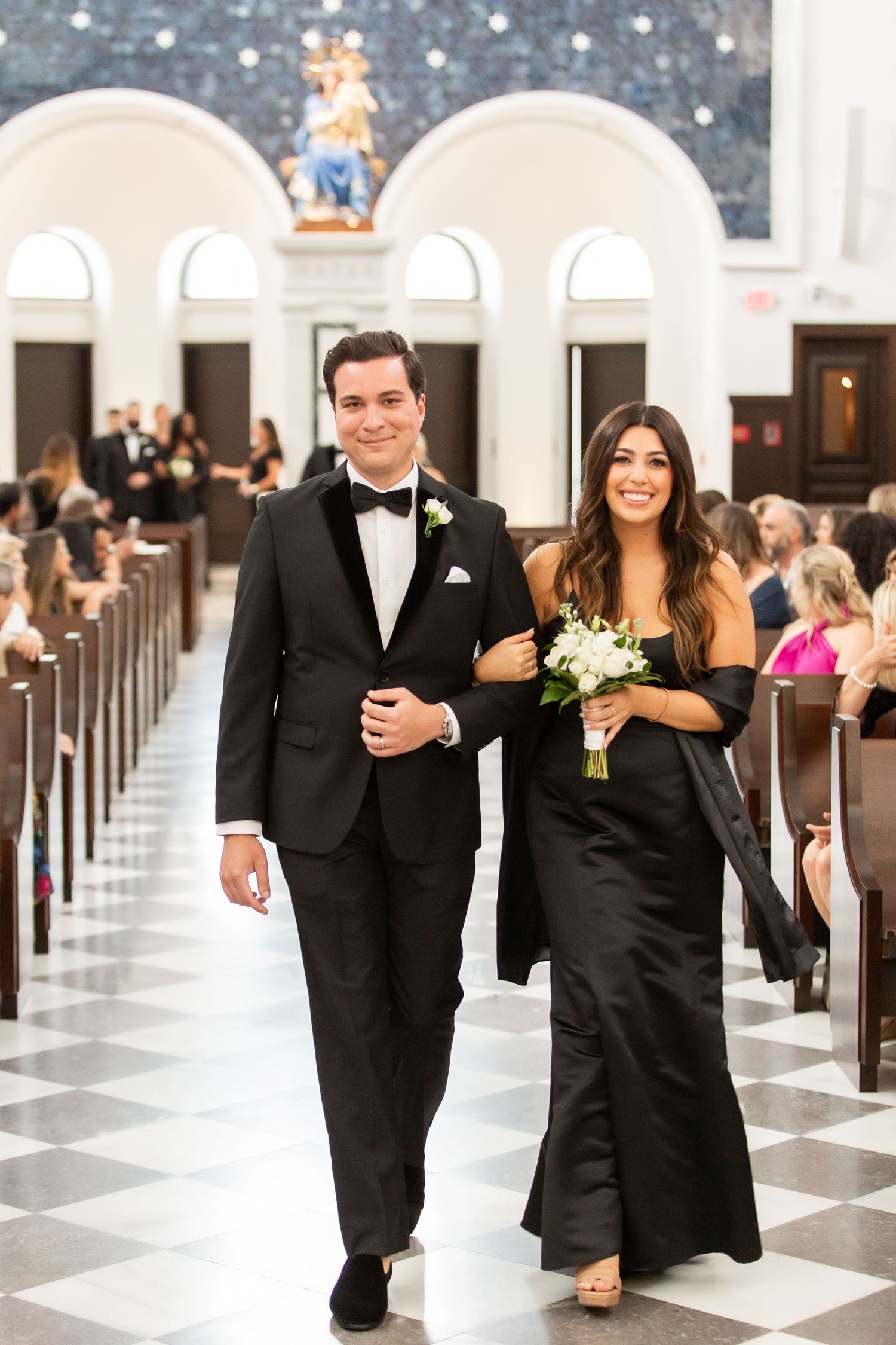 Wedding party members in black attire walking down a church aisle with decorative walls and arched windows.