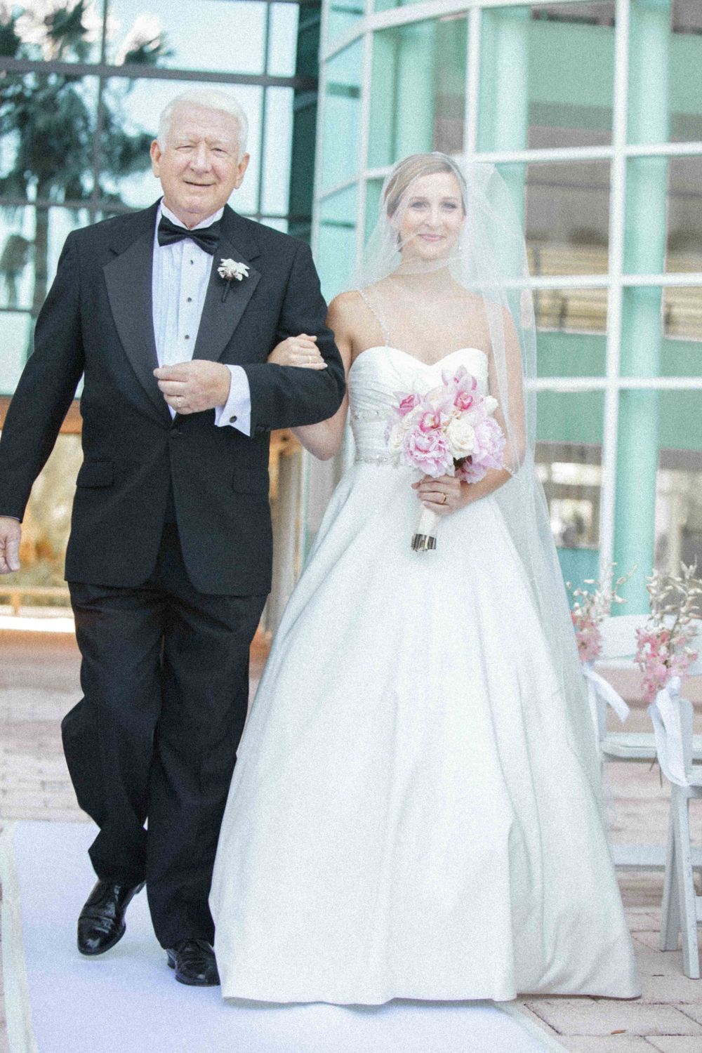 A bride in a white gown holding pink flowers walks down the aisle with a man in a black tuxedo during a wedding ceremony.