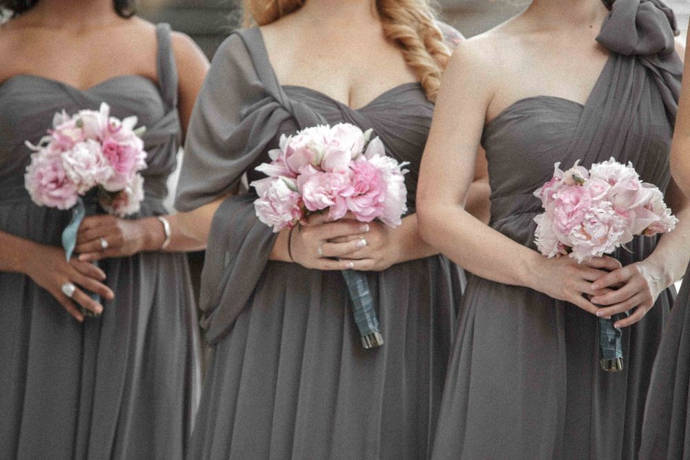 Row of wedding party members in gray dresses holding pink peony bouquets against a gray wall.