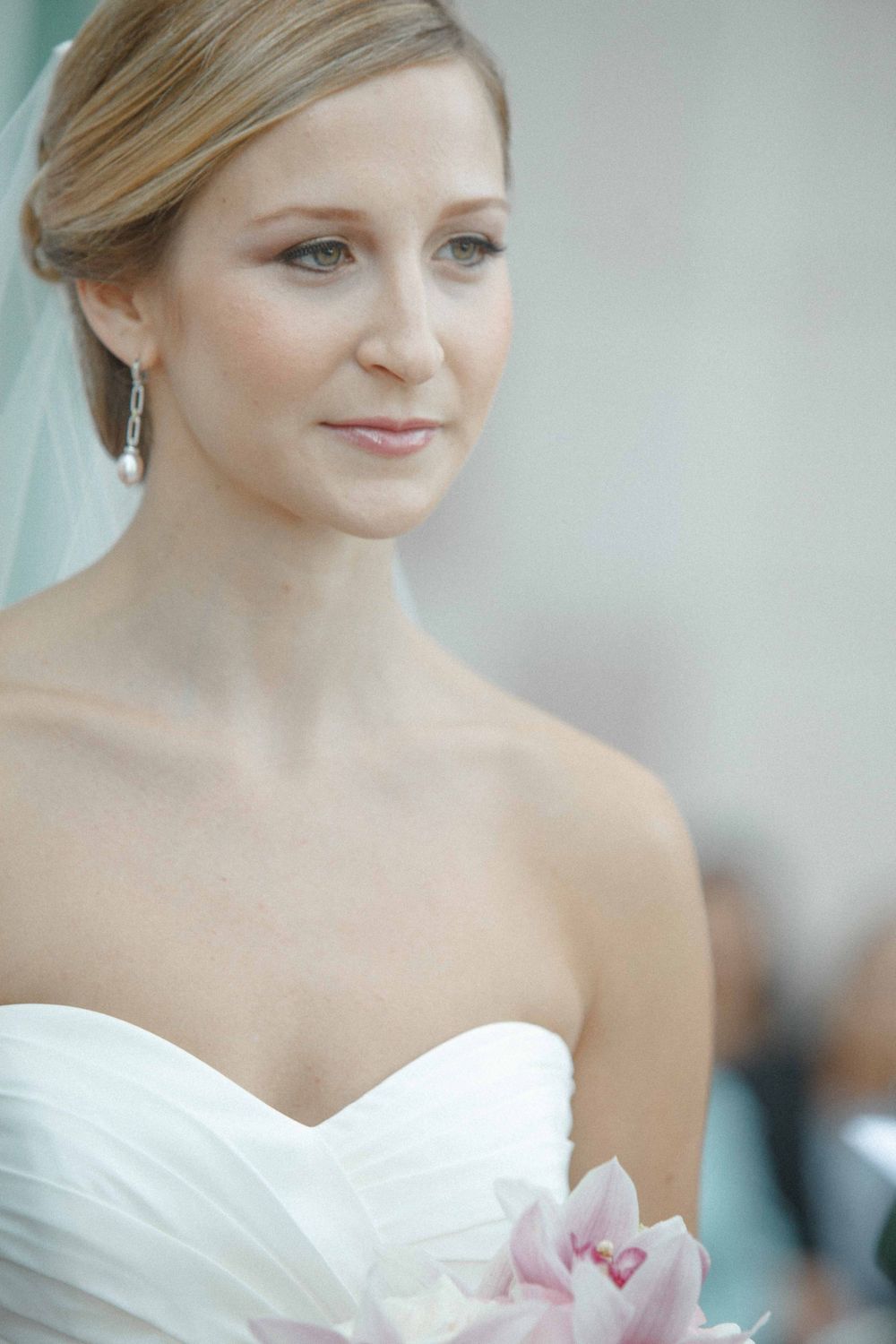 A bride in a strapless white wedding dress holding pink flowers gazes softly to the side with elegant bridal makeup.