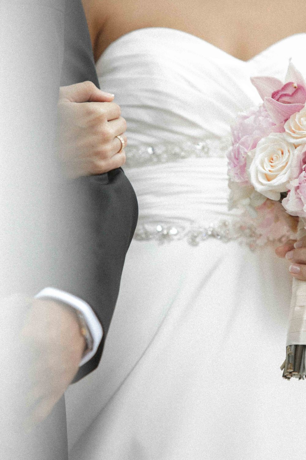 Close-up detail of a white wedding dress bodice and pink rose bouquet held by bride in soft dreamy lighting.