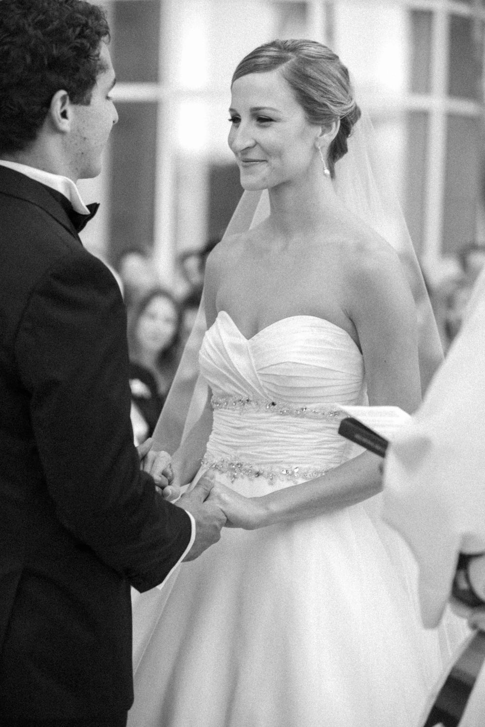 A bride in a strapless white gown gazes lovingly at her groom during their wedding ceremony.