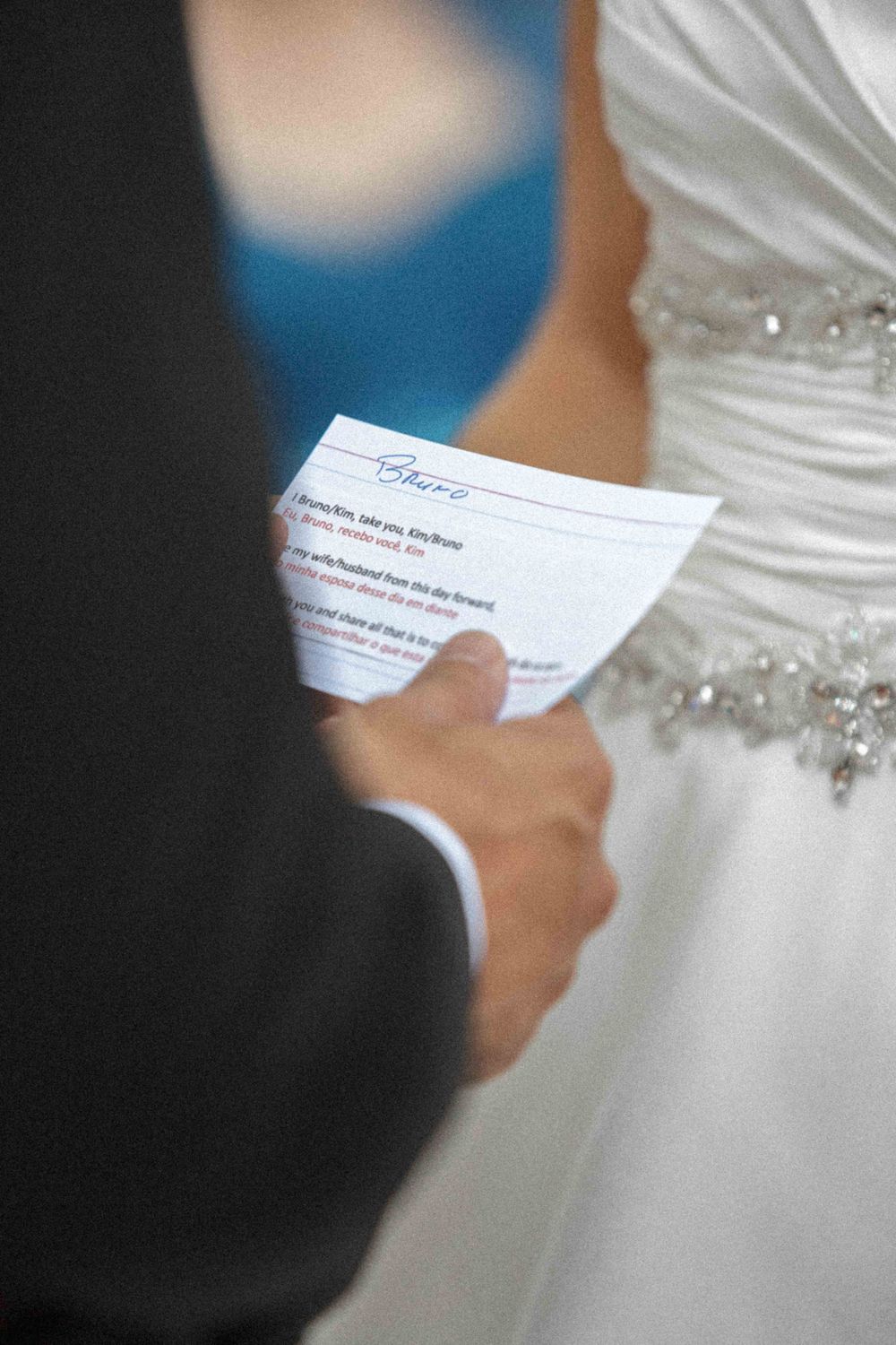Close-up view of hands holding wedding vows during a ceremony with white dress details visible in background.