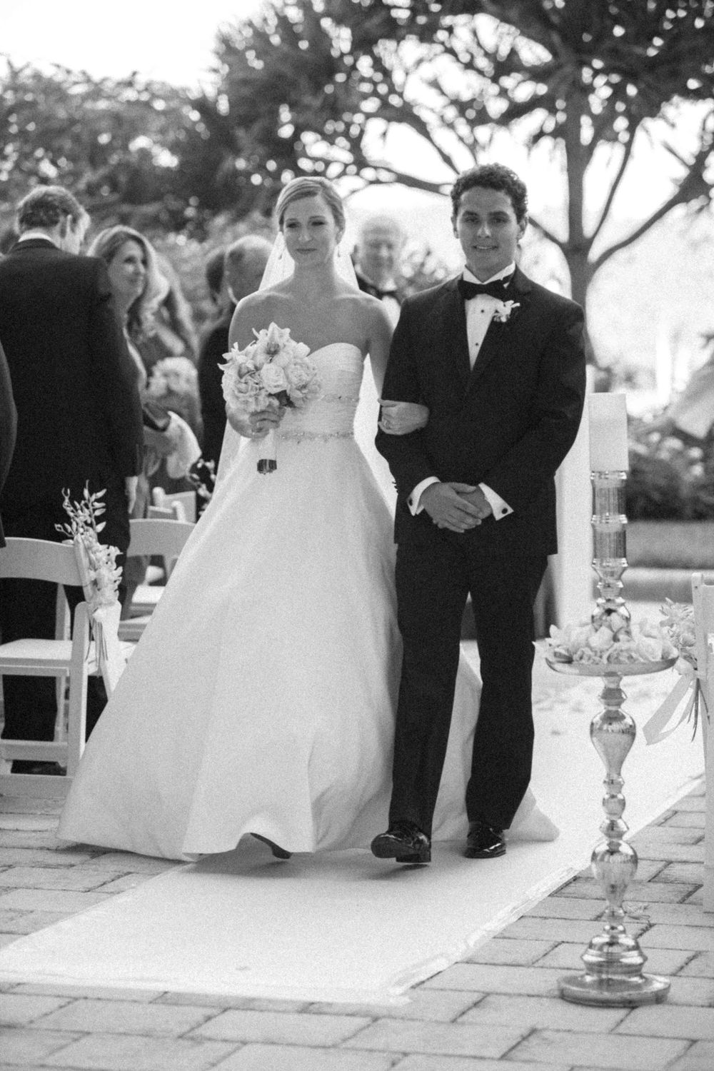 A black and white wedding ceremony photo showing a couple walking down a white aisle runner at an outdoor venue.