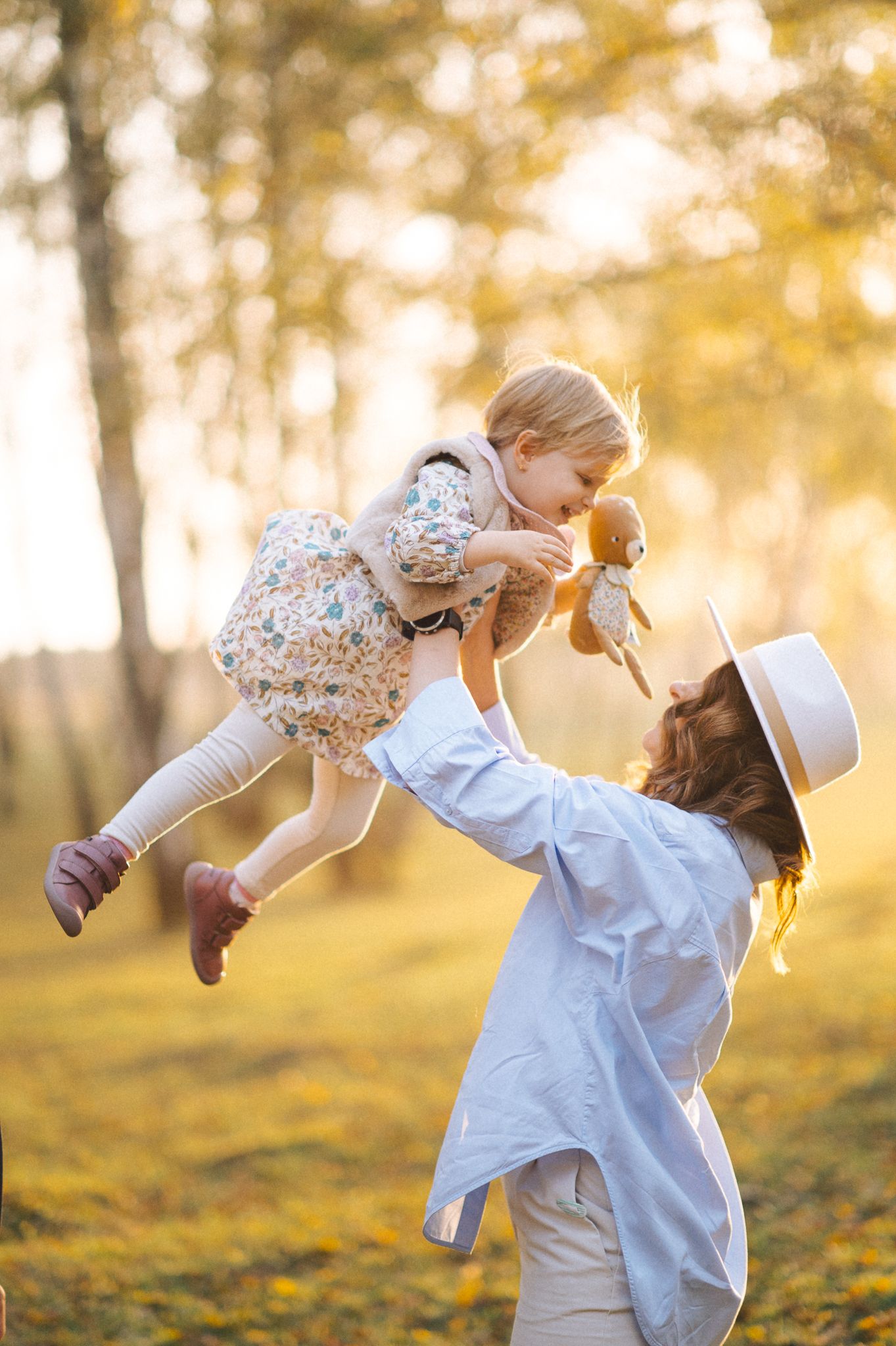 A joyful moment captured in golden sunlight as a figure in white lifts a child wearing a floral dress outdoors.