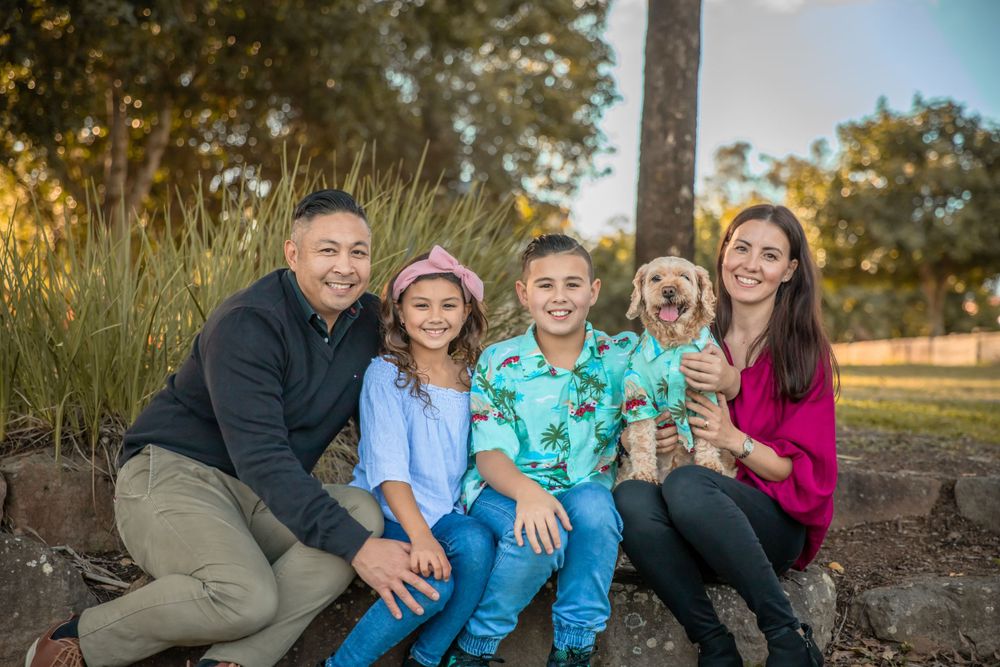 A family of four sit on a stone wall. The young boy and their elderly family dog wear matching blue hawiian shirts