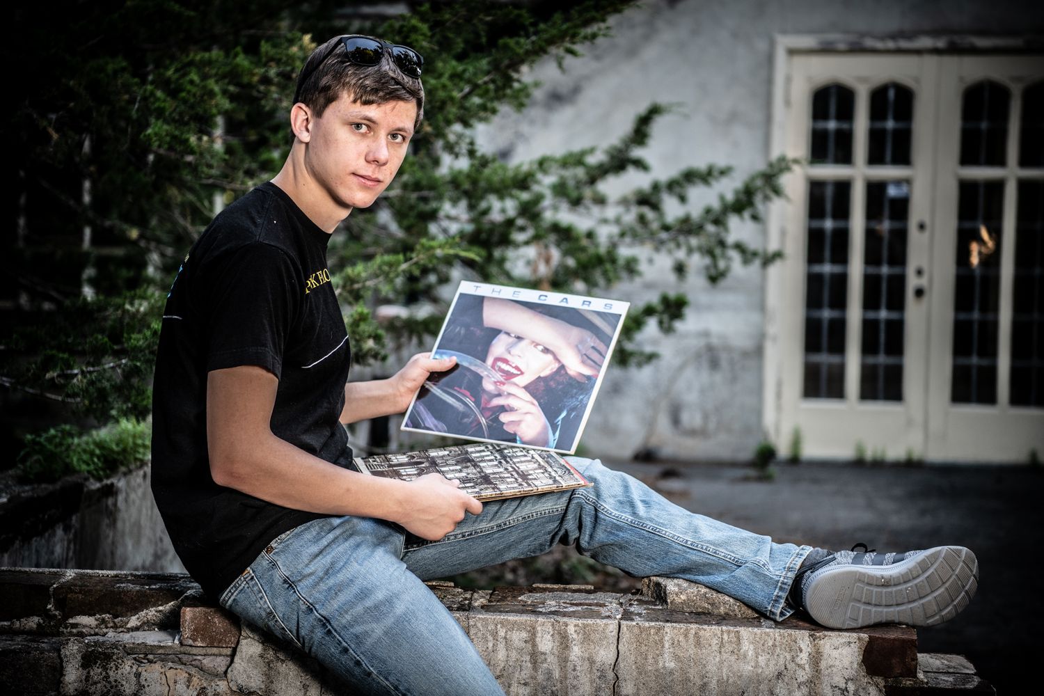Person in black t-shirt sitting outdoors holding laptop with artistic architecture in background.