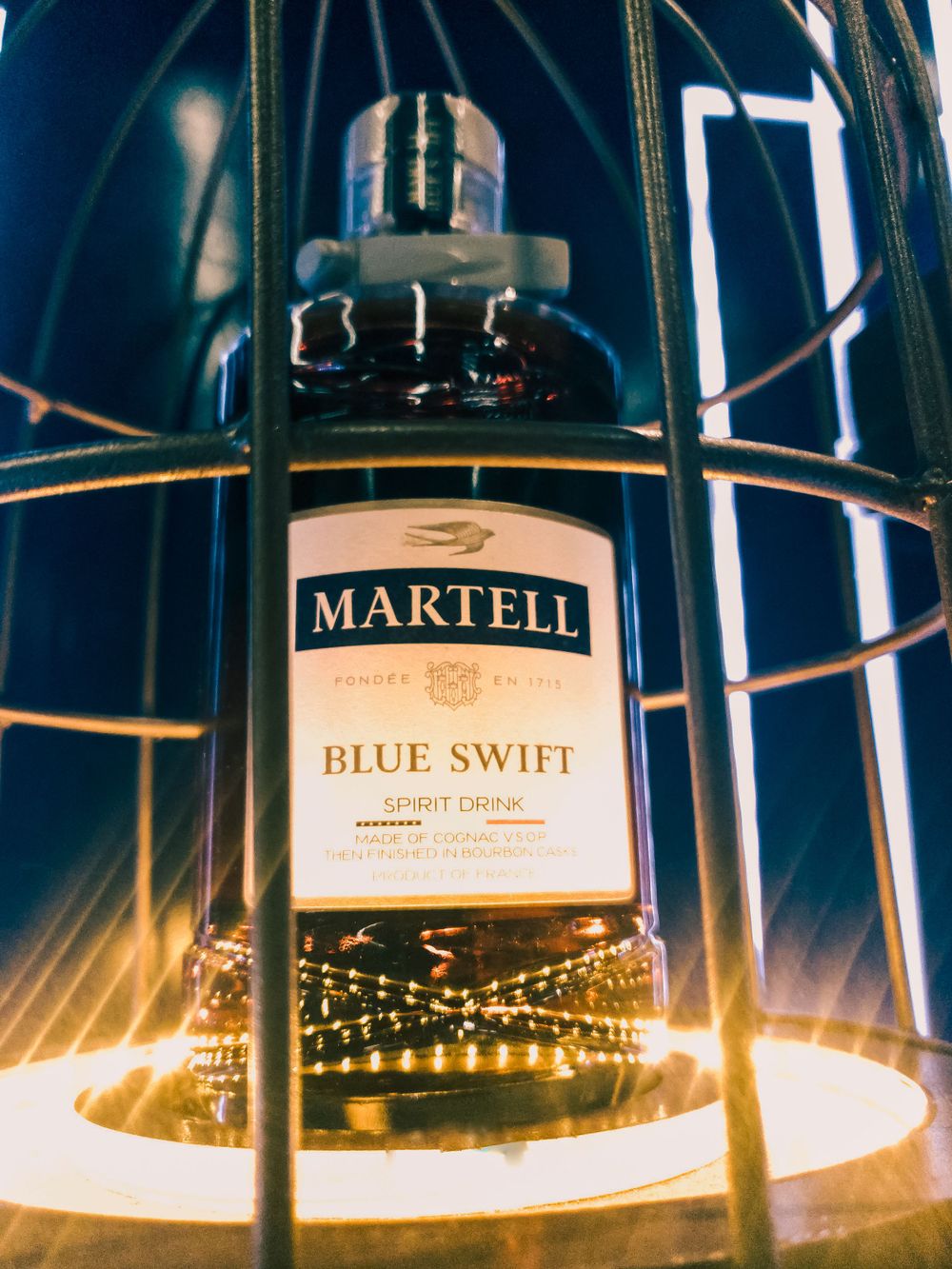 Artistically lit Martell Blue Swift cognac bottle in a display case with dramatic lighting effects.
