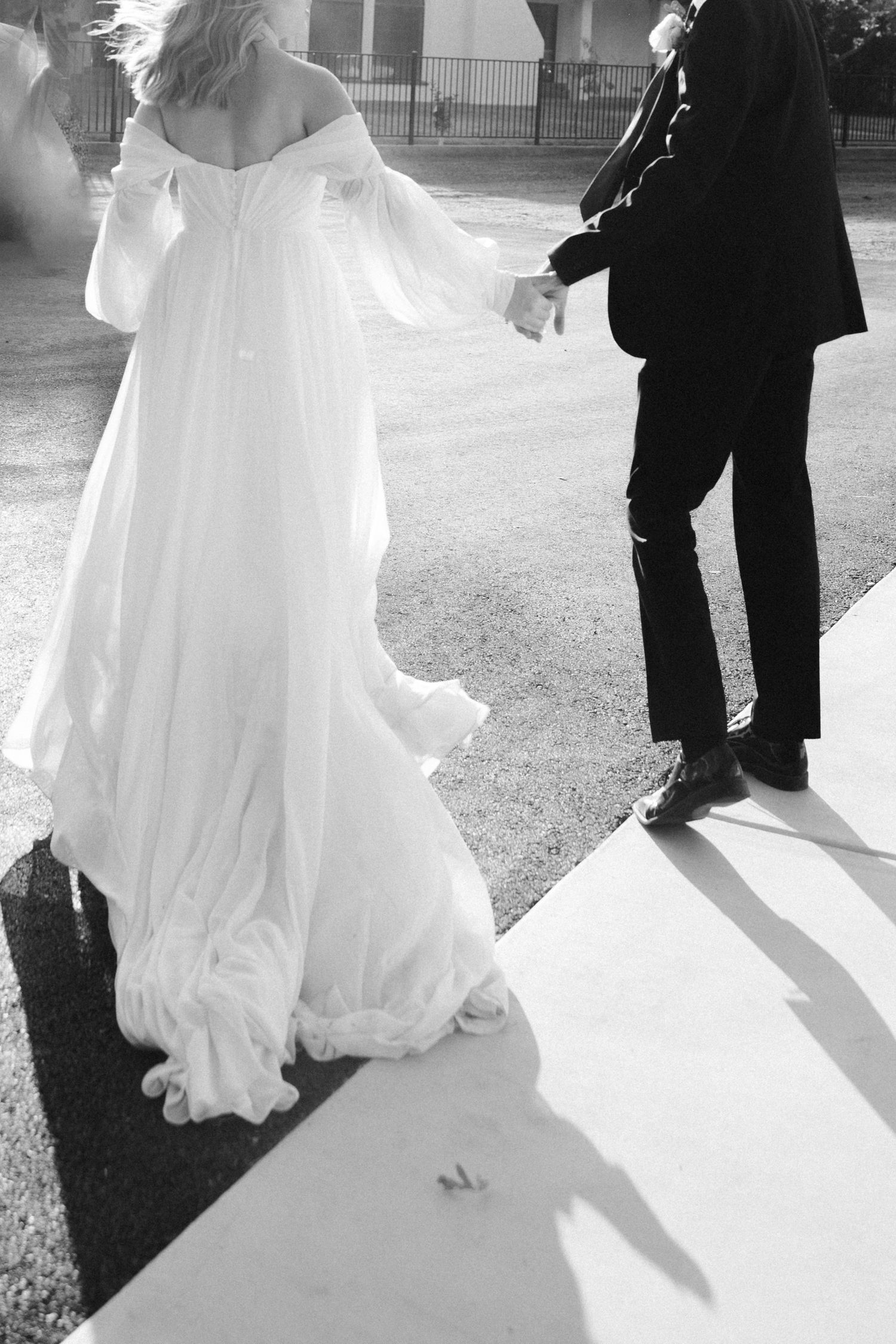 Black and white photo of bride in flowing dress holding hands with groom on sunlit path.