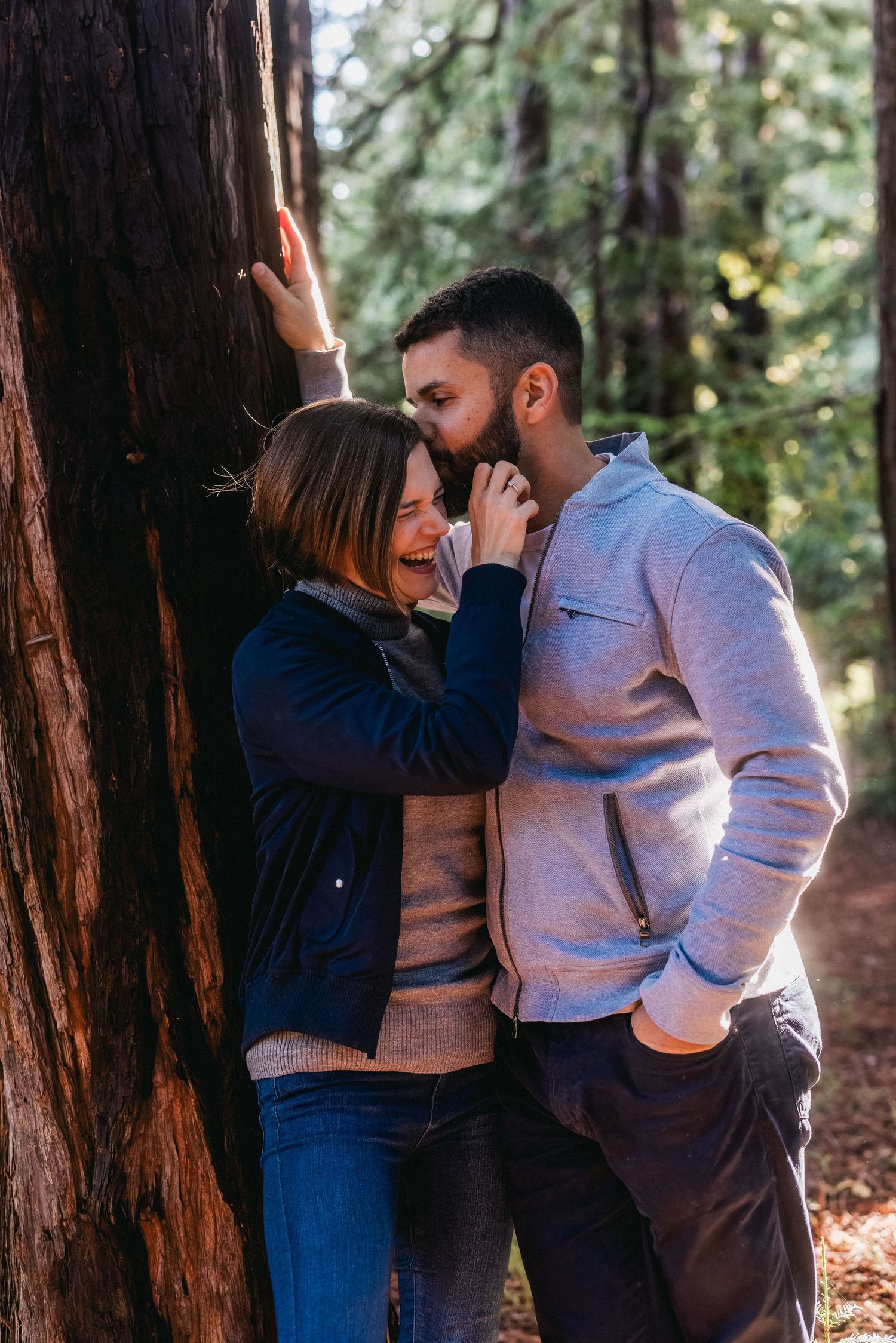 An intimate couple embraces against a redwood tree trunk in a peaceful forest setting.