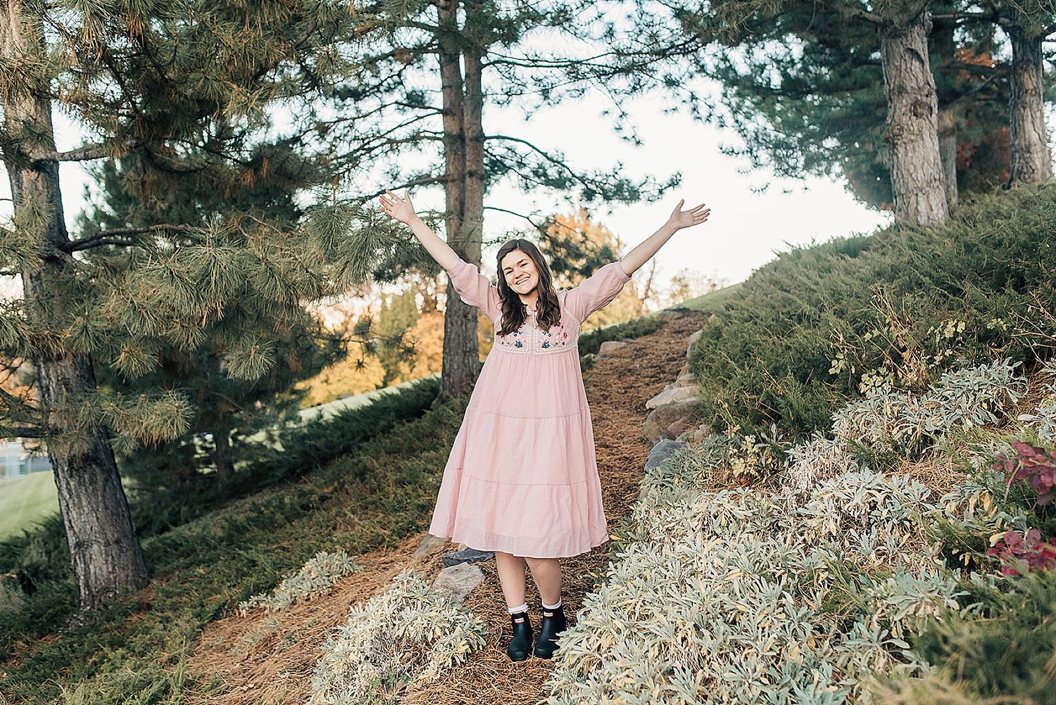 A person in a pink dress poses joyfully with raised arms on a scenic hillside path surrounded by pine trees.