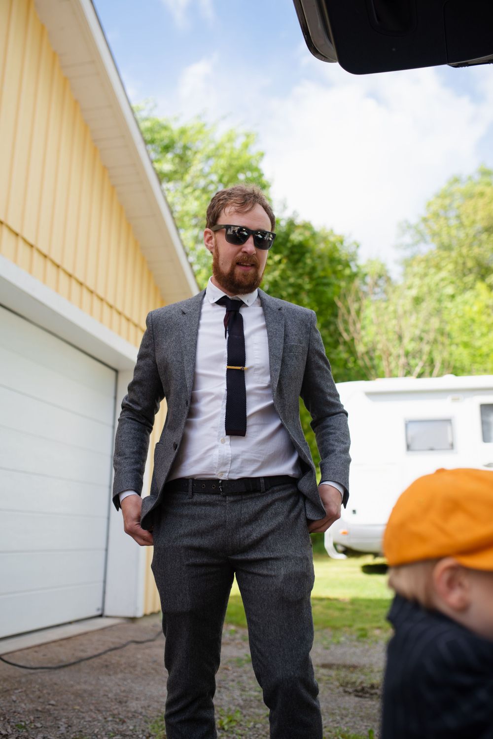 Person in a grey suit standing confidently outside a yellow building on a sunny day.