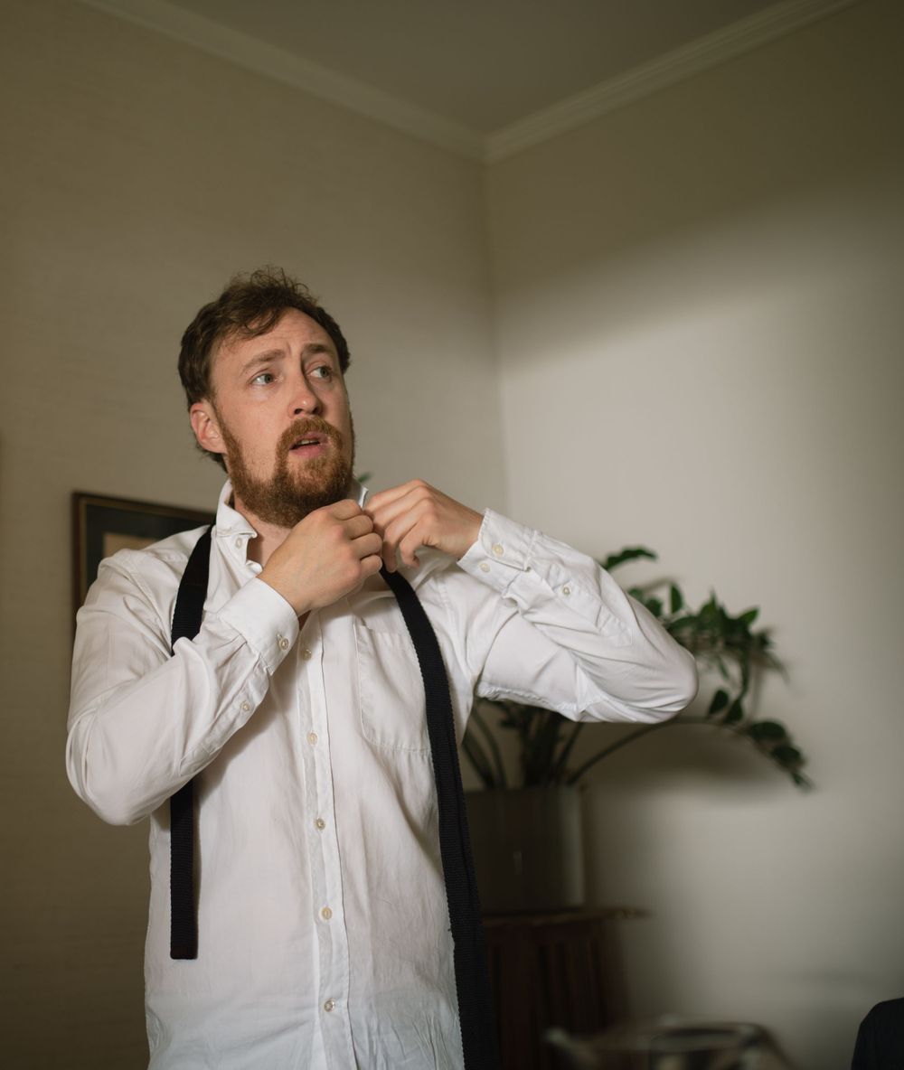 Getting dressed in a formal white shirt and black suspenders in a corner room with a houseplant.