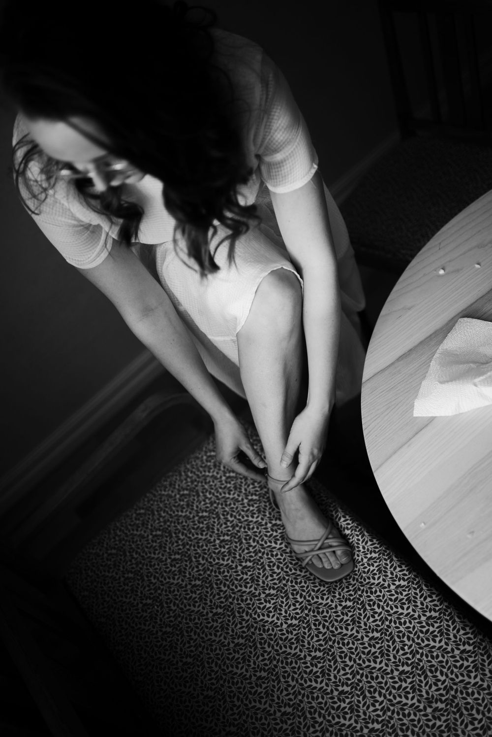Black and white photo of someone fastening sandals while seated on a white chair.