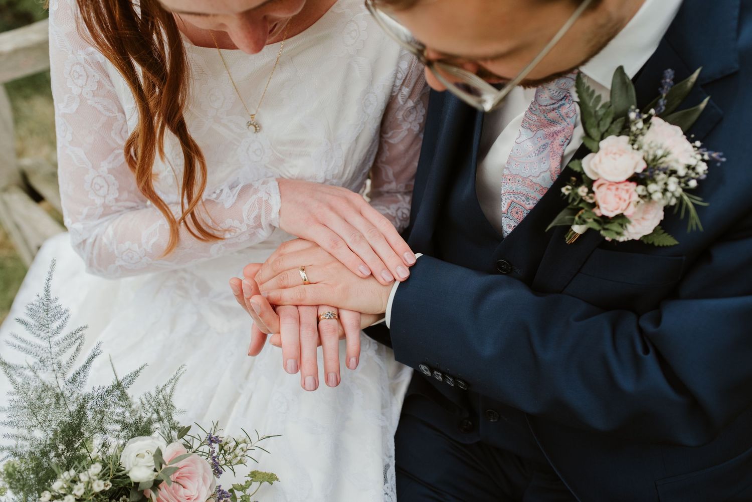 Close-up of newlyweds' hands with rings against white dress and floral bouquet.